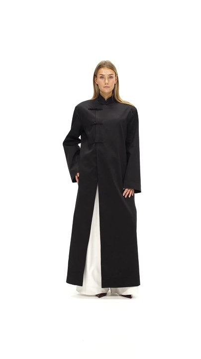 Model walking in the Asymmetric Long Coat in black, worn open over white trousers